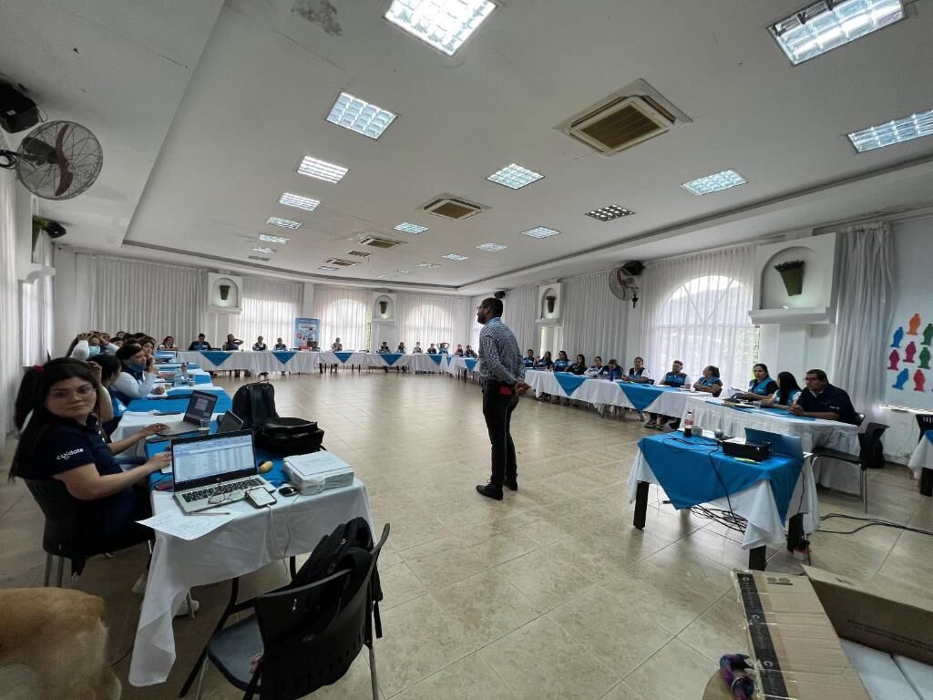 Un salón de conferencias con mesas dispuestas en forma de U, un orador dirigiendo a la audiencia y asistentes sentados en las mesas.