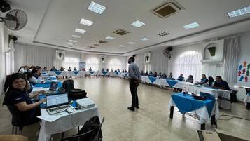 Un salón de conferencias con mesas dispuestas en forma de U, un orador dirigiendo a la audiencia y asistentes sentados en las mesas.