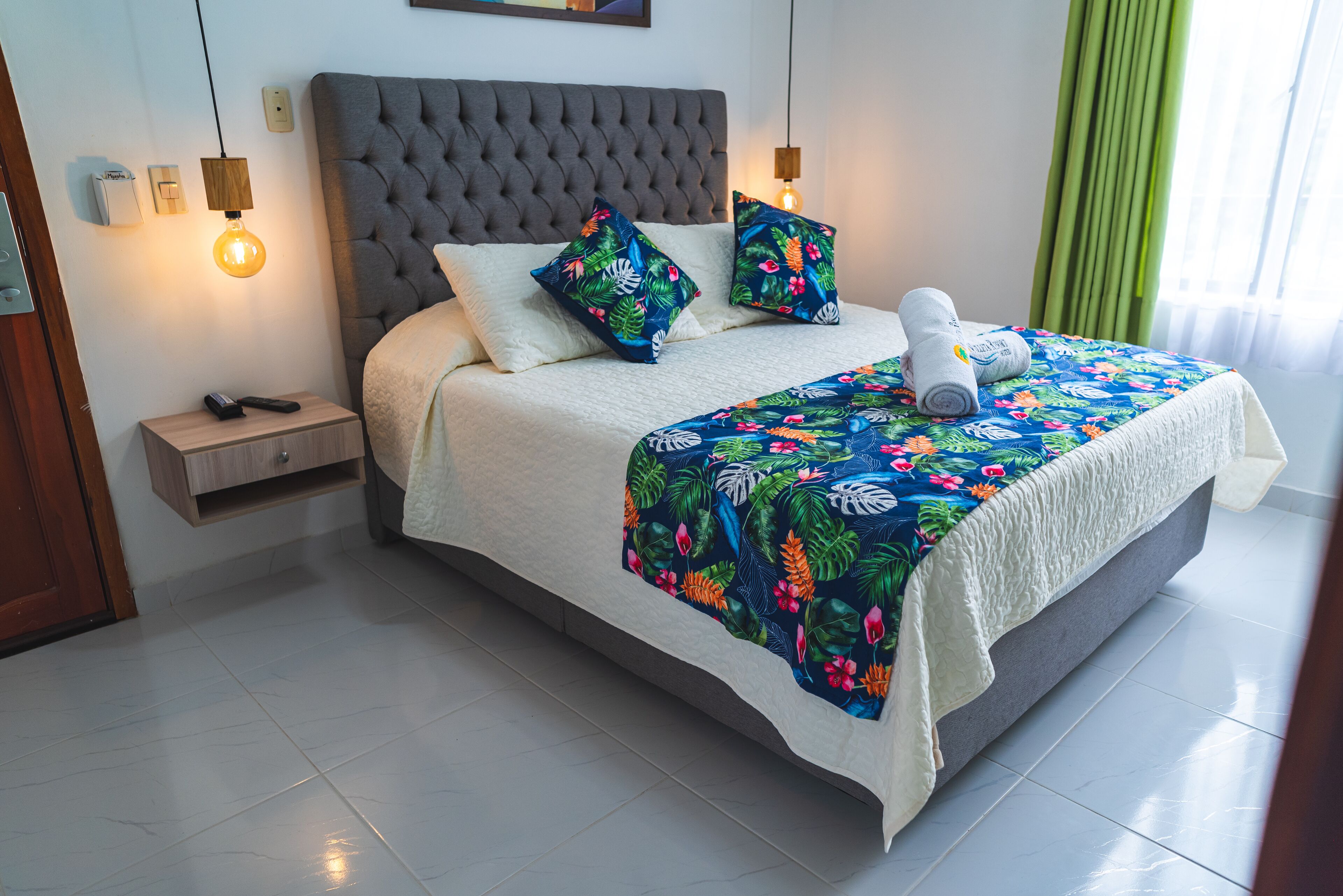 Un lit bien fait, recouvert d’une couvre-lit à motifs floraux, avec une tête de lit rembourrée de couleur grise et deux oreillers ornés de motifs tropicaux.