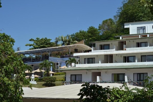Un hotel de varios pisos con tejado plano, balcones y un área exterior cubierta con sombrillas.