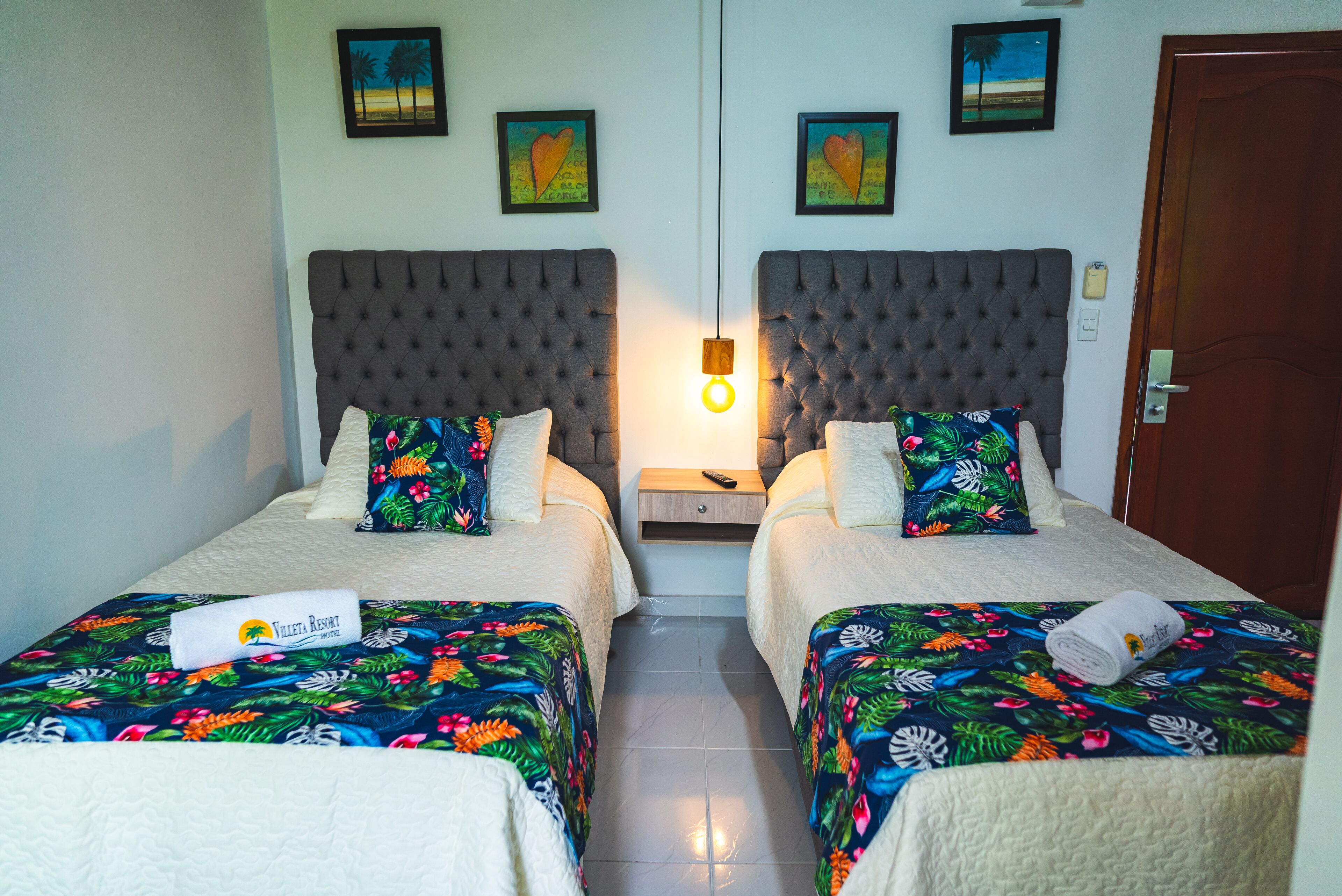 Een hotelkamer met twee bedden, elk voorzien van een bloemenbedekker en een kussen met een tropisch ontwerp. Tussen de bedden staat een nachtkastje met een lamp, en aan de rechterkant een deur.