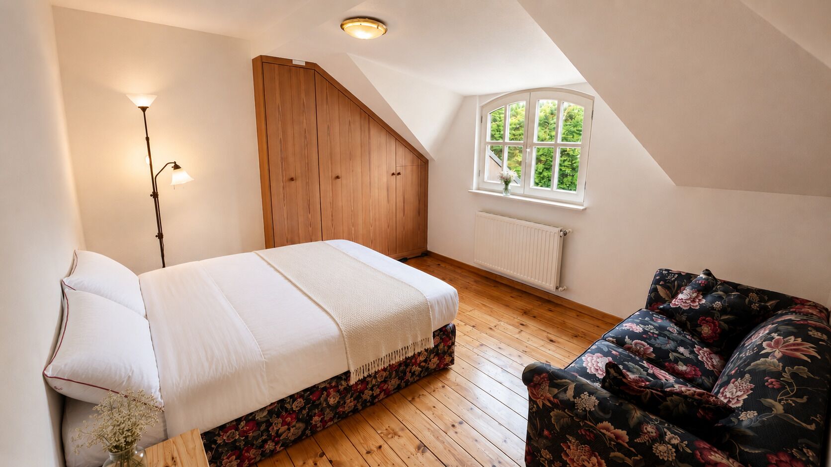 Ein Schlafzimmer mit einem Bett, einem Nachttisch, einer Stehlampe, einem Fenster mit Blick und einem floralen Sofa.