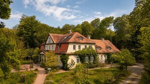 A two-story house with a red-tiled roof, surrounded by lush greenery and a gravel path leading to the entrance. - Seegut Blaue Blume (Boitzenburger Land)