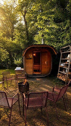 A wooden cabin with a fire pit and metal chairs in a forest setting.