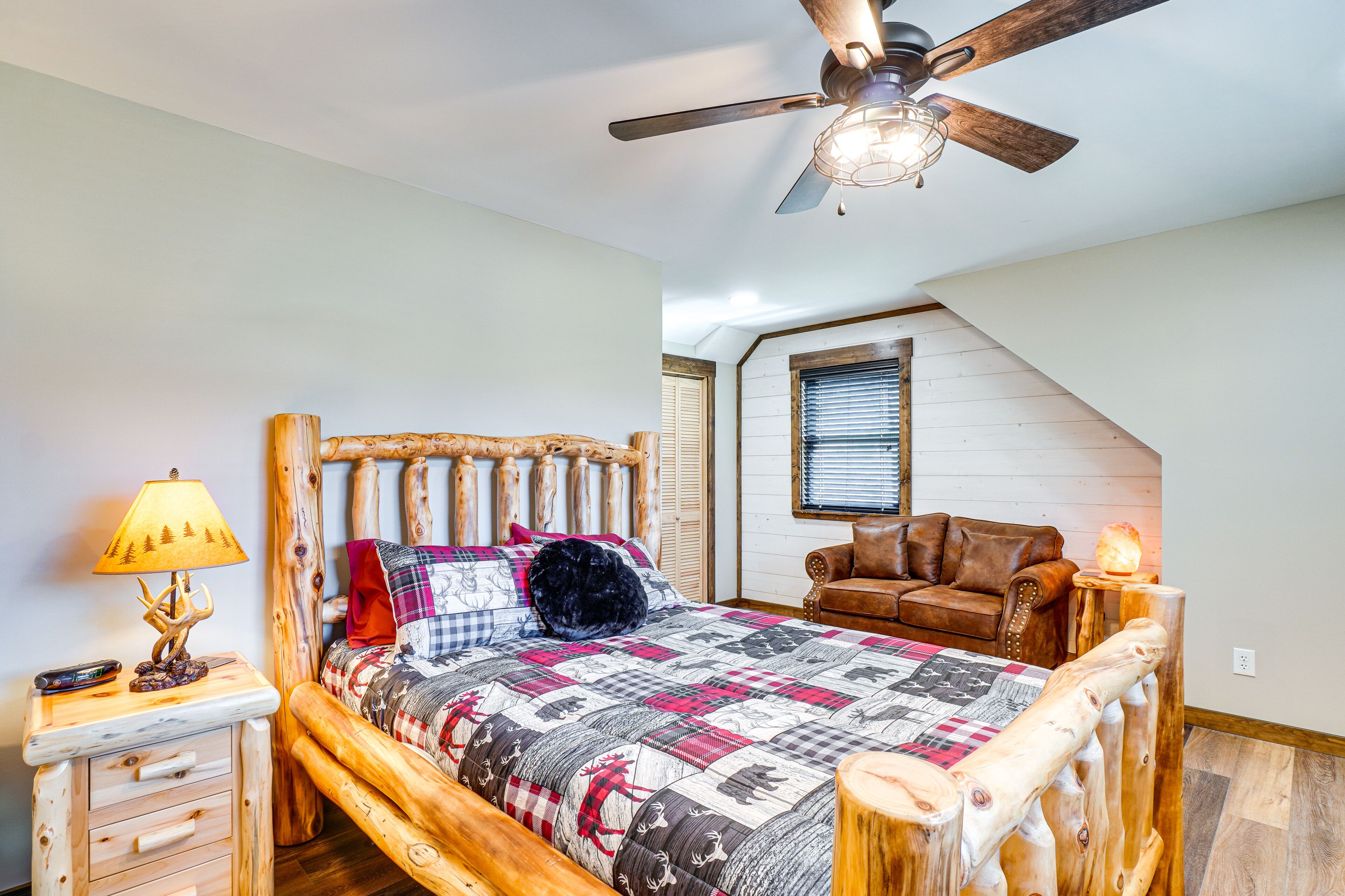 A cozy bedroom with a wooden bed, a nightstand, a lamp, a ceiling fan, and a window with blinds.