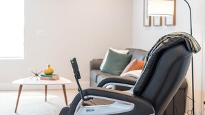 Una silla de masaje en un salón moderno con una pared blanca, una mesita redonda y un sofá con cojines.