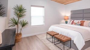 Une chambre à coucher comprenant un lit, deux poufs, une table de chevet, une lampe, une plante en pot et un plafond en bois.