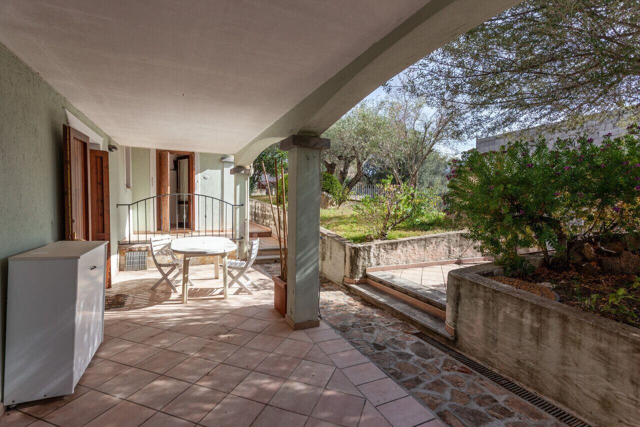 A covered patio area with a table and chairs, a refrigerator, and a view of the garden and trees.