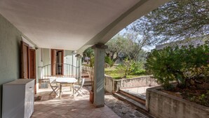 Ein überdachter Patio-Bereich mit Tisch und Stühlen, einem Kühlschrank sowie Blick auf Garten und Bäume.