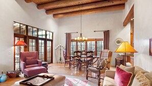 A spacious living room with a high ceiling, wooden beams, a dining area with a table and chairs, and a large window providing natural light. - Casa Segunda | Elegant Santa Fe Retreat (Santa Fe)