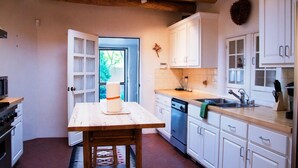 A kitchen with white cabinets, a wooden table, and a view of greenery outside. - Casa del Monte Sol | Charming Adobe Retreat (Santa Fe)