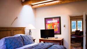 A bedroom with a bed, a television on a wooden table, a painting on the wall, and a view of a room with a sofa and a window. - Casa del Monte Sol | Charming Adobe Retreat (Santa Fe)