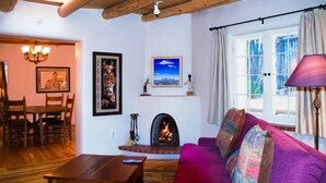 A cozy living room with a fireplace, a sofa, a dining table, and a chandelier. - Casa del Monte Sol | Charming Adobe Retreat (Santa Fe)