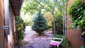 A narrow outdoor pathway leading to a bench and a table, surrounded by greenery and a large evergreen tree. - Casa del Monte Sol | Charming Adobe Retreat (Santa Fe)