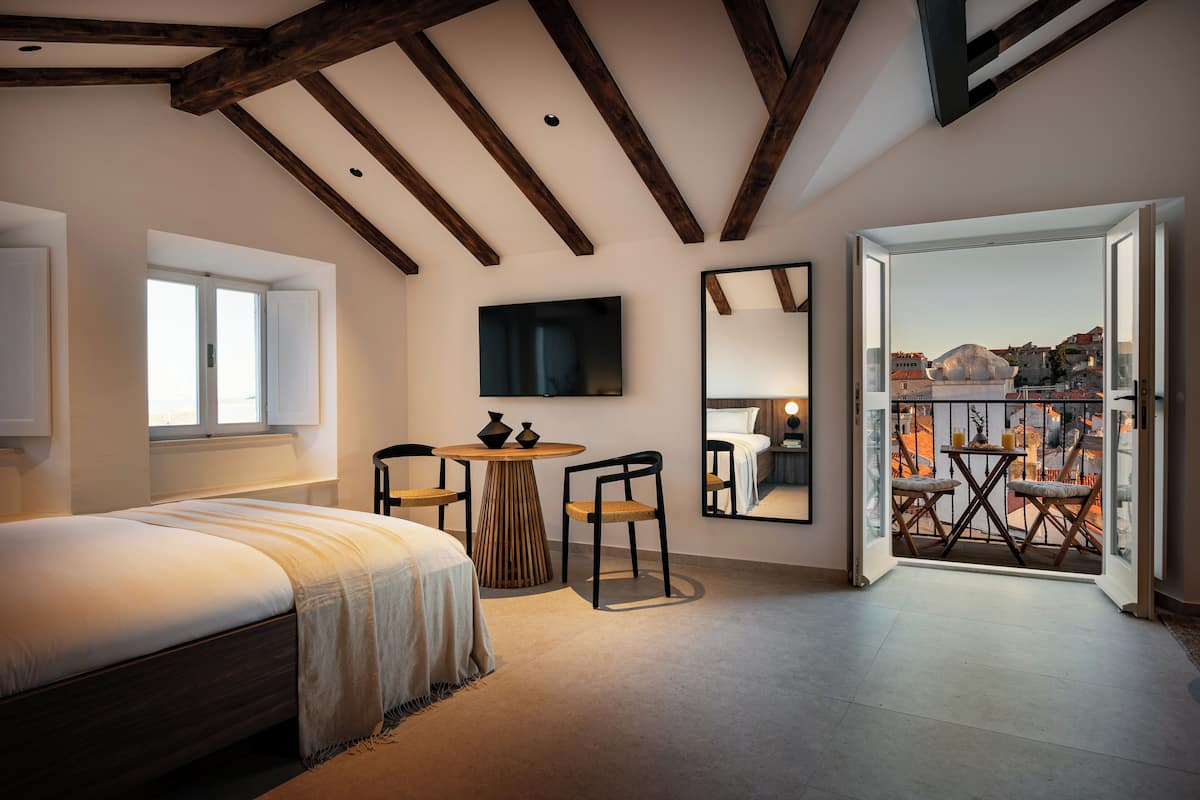 Un dormitorio con cama, una mesa redonda de madera, dos sillas, televisor, un espejo grande y un balcón con vistas a la ciudad.