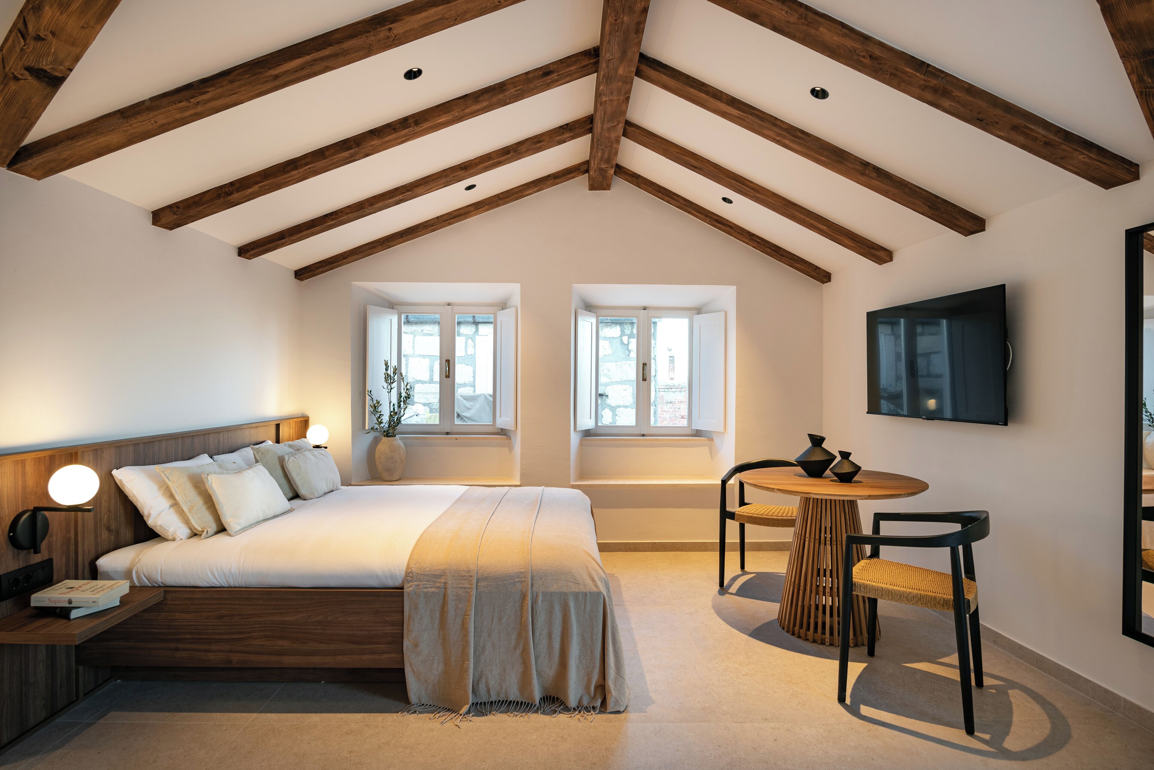 Un dormitorio con techo de madera, una cama con cabecera, dos ventanas, una mesita de noche, una silla y un televisor montado en la pared.