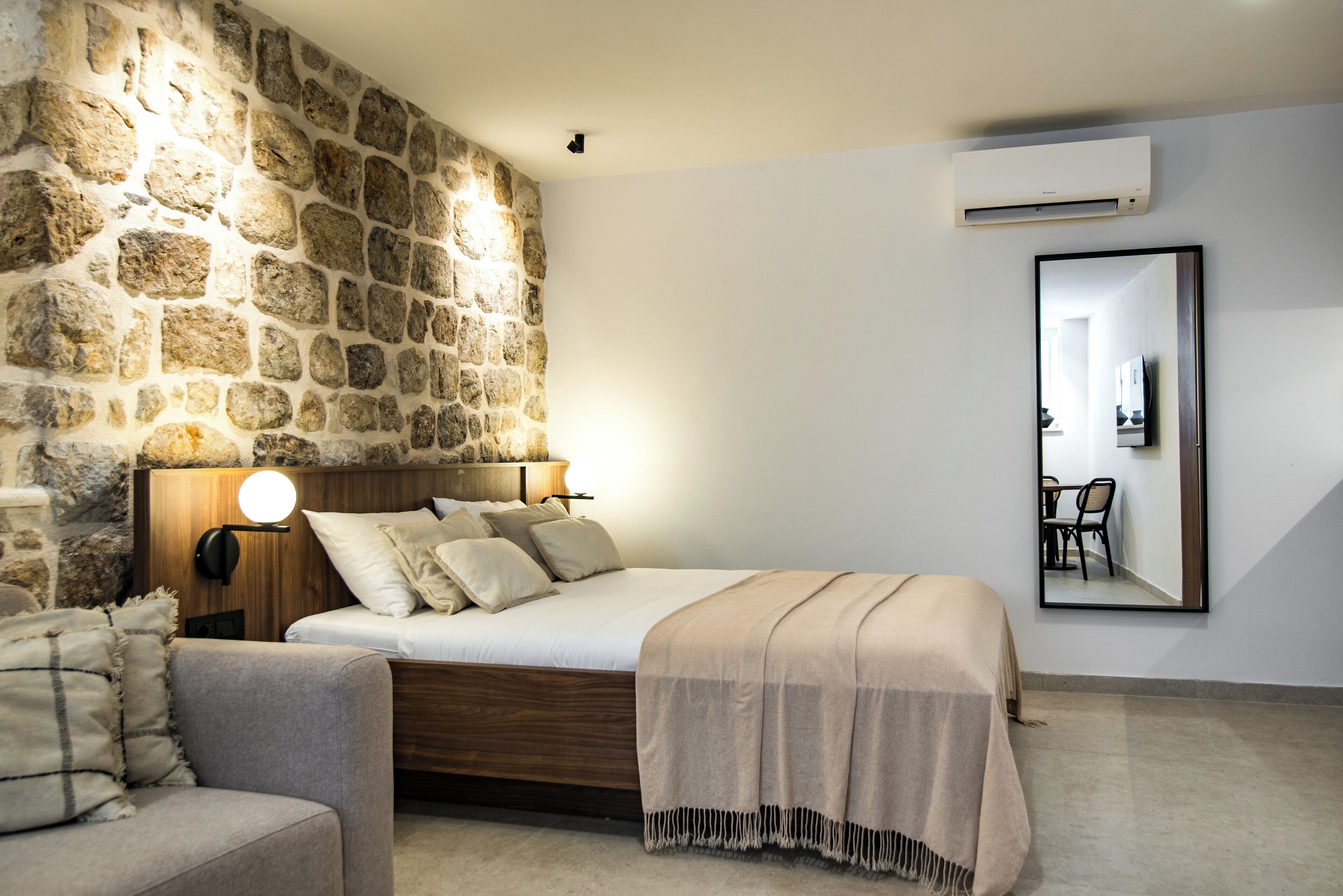 A bedroom with a stone wall, a bed with a wooden headboard, a bedside table with a lamp, a chair, and a mirror on the wall.