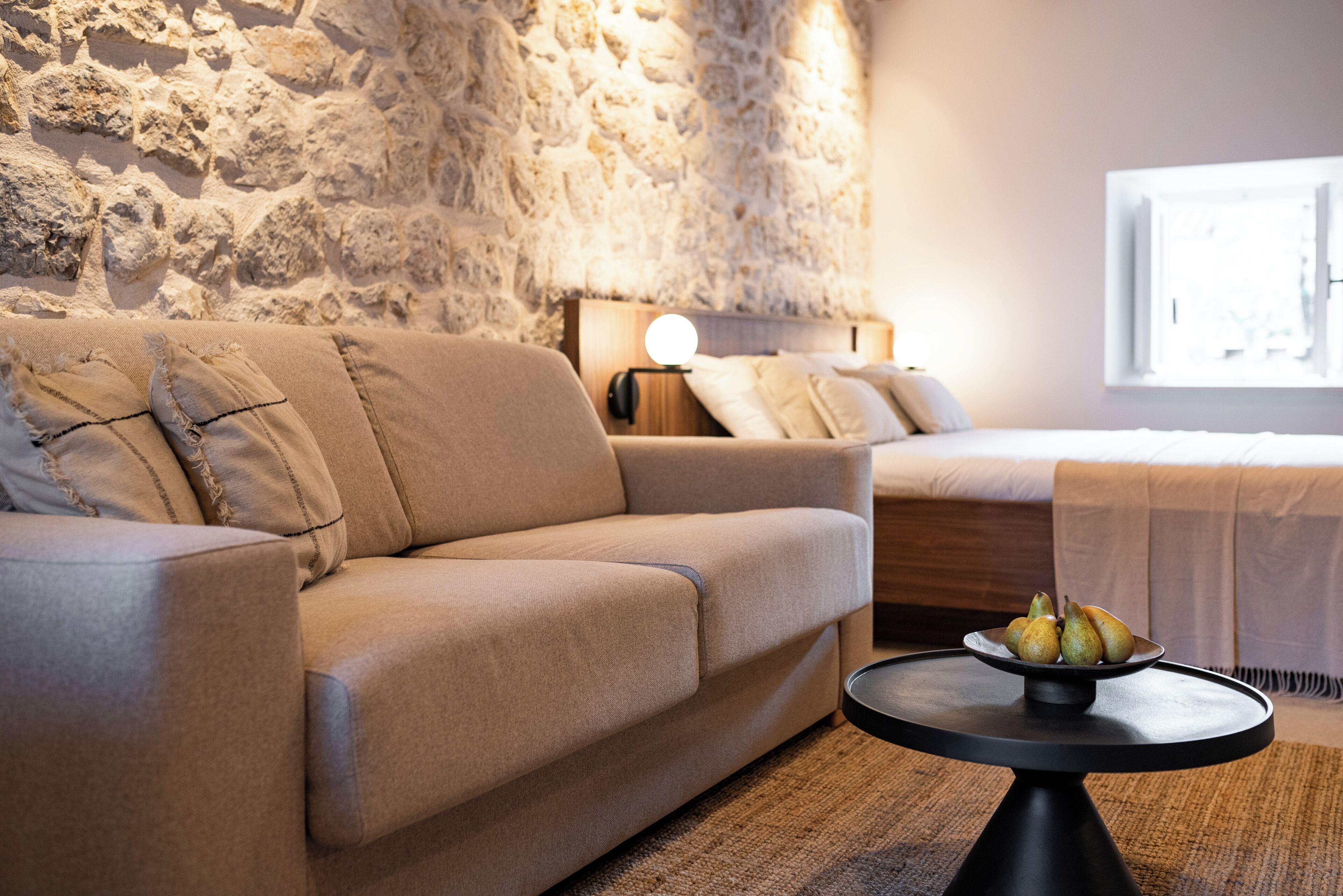 Una sala moderna con una pared de piedra, un sofá, una mesita de noche con una lámpara y una cama con cabecera blanca.