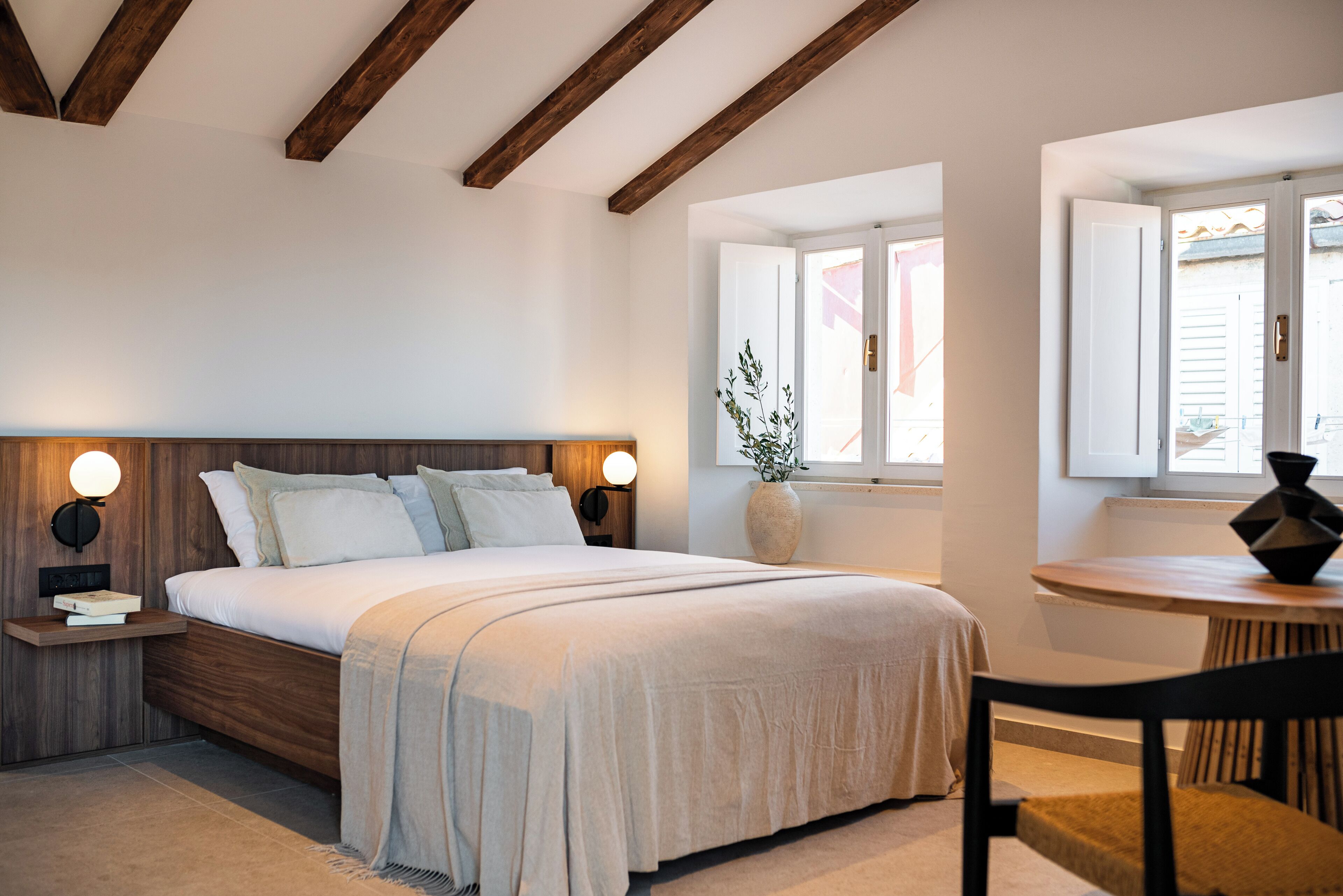 A bedroom with a wooden headboard, a bedside table with a lamp, a large bed with white bedding, a window with blinds, and a small table with a chair.