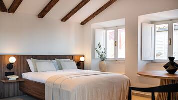 Un dormitorio con cabecera de madera, mesita de noche con lámpara, cama grande con ropa blanca, ventana con persianas y una pequeña mesa con una silla.