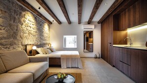 A modern bedroom with a stone wall, wooden ceiling beams, a bed, a bedside table, and a kitchen area with wooden cabinets.