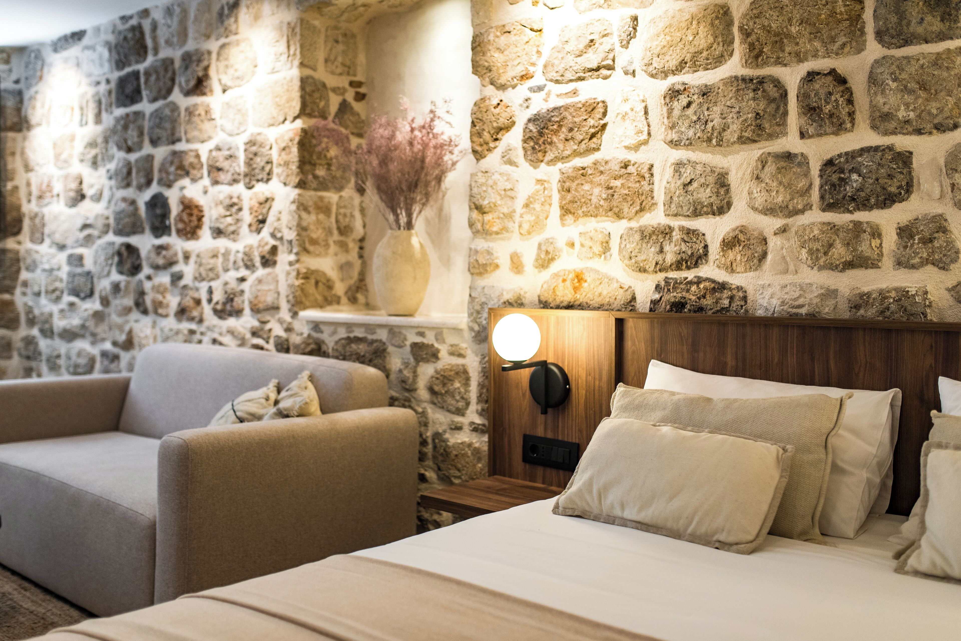 A room with a stone wall, a bed with beige bedding, a wooden headboard, a nightstand, a lamp, and a vase with dried flowers.