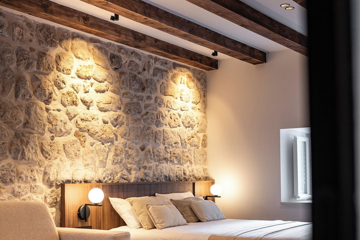 Un dormitorio con pared de piedra, vigas de madera, cama con cabecera, lámparas de noche, sofá y una mesa redonda con fruta.