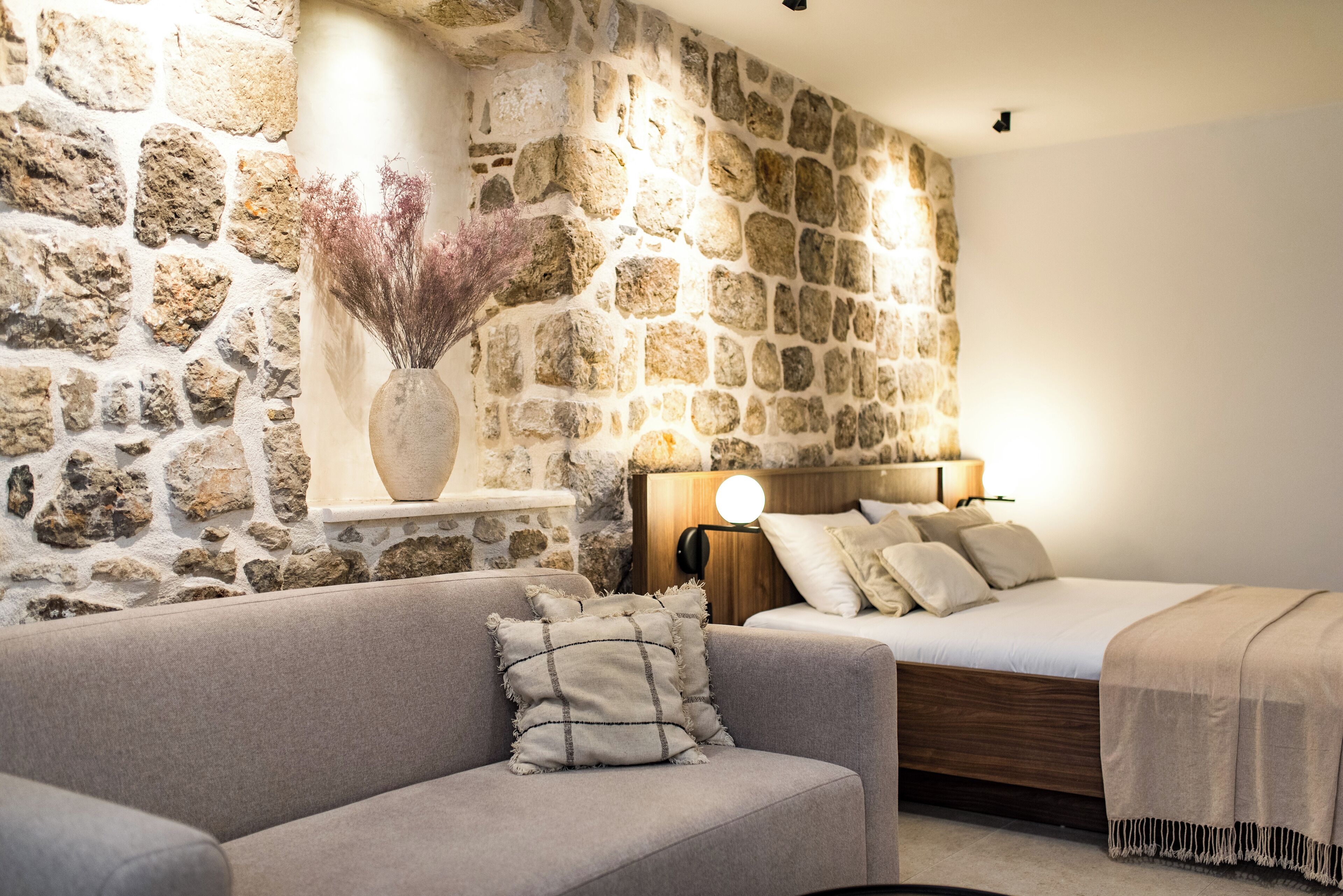 Quarto com parede de pedra, cama de madeira, sofá cinza e mesa de cabeceira com abajur.