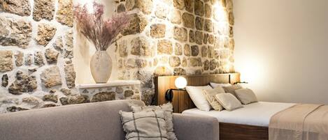 Quarto com parede de pedra, cama de madeira, sofá cinza e mesa de cabeceira com abajur.