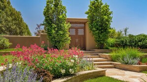 A well-maintained garden with a variety of flowers and shrubs, a wooden door, and a stone pathway leading to a building with a beige facade. - Villa Campanas | Luxury Home with Mountain Views (Santa Fe)