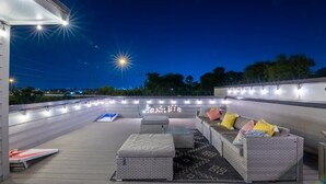 An outdoor deck with a sofa and ottoman, string lights, and a view of the city at dusk. - Two New Lux Homes! Rooftop + Hot Tub, Pool & Sauna (Nashville)