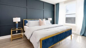 A modern bedroom with a dark blue accent wall, a wooden bed frame, and a large window with curtains. - 2 Lux Homes, 8 bedrooms/28 beds! Baller Game Room (Nashville)