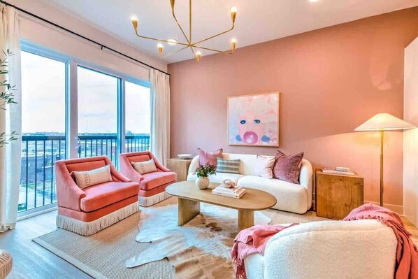 A modern living room with a large window, a white sofa, a pink armchair, and a coffee table.
