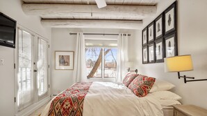 Een slaapkamer met een groot bed, een nachtkastje, een lamp, een raam met gordijnen, een ingelijst kunstwerk aan de muur en een deur met luiken.