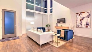 Ein modernes Wohnzimmer mit einem weißen Sofa, blauen Ziertelementen, einem kleinen runden Tisch und einem blauen Sessel. Es gibt ein großes Fenster mit Jalousien, einen flachbildfernseher an der Wand und ein abstraktes Kunstwerk.