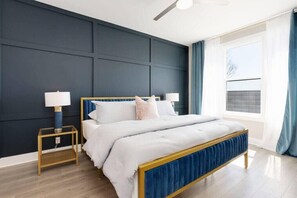 A modern bedroom with a dark blue accent wall, a wooden bed frame, and a large window with curtains.