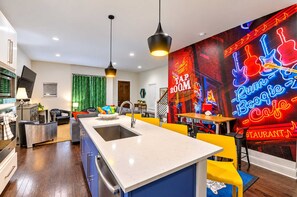 A modern kitchen with a central island, pendant lighting, and a vibrant neon mural. - Lux Designer Gulch Home - Skyline Views - 11 Beds! (Nashville)