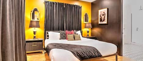 Un dormitorio con una cama, dos mesitas de noche con lámparas, un ventilador de techo, una puerta y un cuadro en la pared.