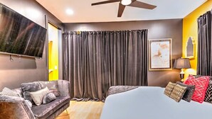 Een slaapkamer met een bed, een bank, een televisie, een plafondventilator en een raam met gordijnen.