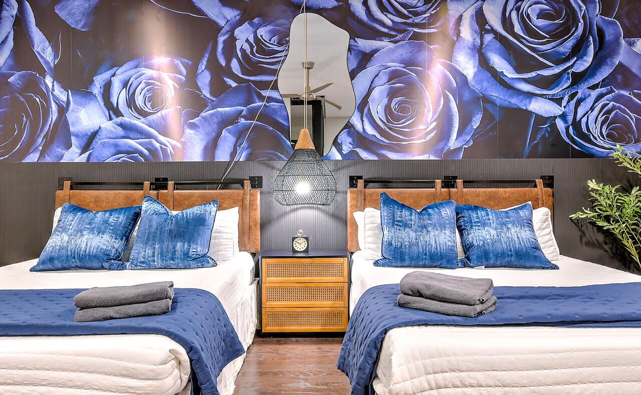 A hotel room with two beds, each with a wooden headboard, blue bedding, and a wall mural of blue roses. There is a small wooden nightstand with a clock between the beds, and a hanging light fixture above.