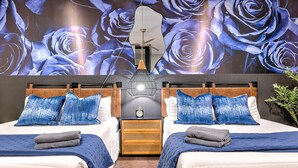 A hotel room with two beds, each with a wooden headboard, blue bedding, and a wall mural of blue roses. There is a small wooden nightstand with a clock between the beds, and a hanging light fixture above. - Premium Gulch Home 15 Beds Ultimate Skyline Views (Nashville)