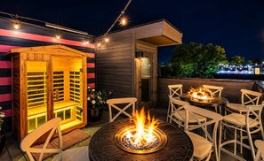 Eine Terrassenausstattung im Freien mit Feuerschale, weißen Stühlen und einer Holzhütte mit Glastür.