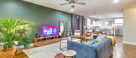 Una sala moderna con un sofá azul, una mesa de centro y un televisor. Se observa una cocina al fondo con un refrigerador y un comedor.
