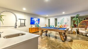 A modern living room with a pool table, a large flat-screen TV, a sink, and a decorative plant.