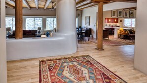 A spacious living area with a high ceiling, exposed wooden beams, a large rug, and a kitchen in the background. - Casa San Miguel | Santa Fe Sanctuary with Views (Santa Fe)