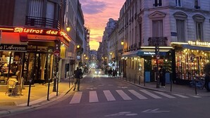 A street view at dusk with buildings on either side, a pedestrian crossing, and storefronts including a restaurant and a bar. - Charming Apartment in the Heart of Paris. Families are Welcome (Paris)