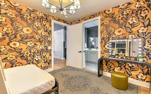 A bedroom with floral wallpaper, a bed, a vanity with a mirror and lights, and a door leading to a bathroom.