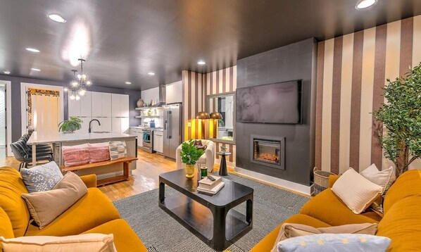 A modern living room with a sofa, coffee table, and fireplace. There is a kitchen area with cabinets and a dining table.