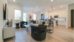 Amplia sala de estar con sofá, mesa de centro y televisor sobre un mueble bajo. Se aprecia una cocina al fondo con alacenas y microondas.