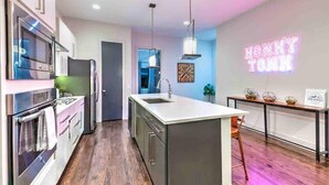 A modern kitchen with stainless steel appliances, a central island, and a neon sign on the wall. - Premiere Gulch Home, 15 Beds! Best Skyline Views! (Nashville)